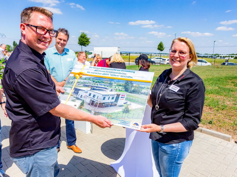 Ein Mann und eine Frau halten gemeinsam eine gerahmte Darstellung eines neuen Firmengebäudes