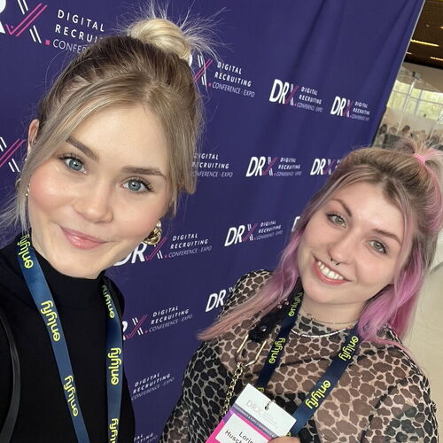 Zwei junge Frauen mit Namensschildern und Lanyards vor einer blauen Wand mit der Aufschrift \