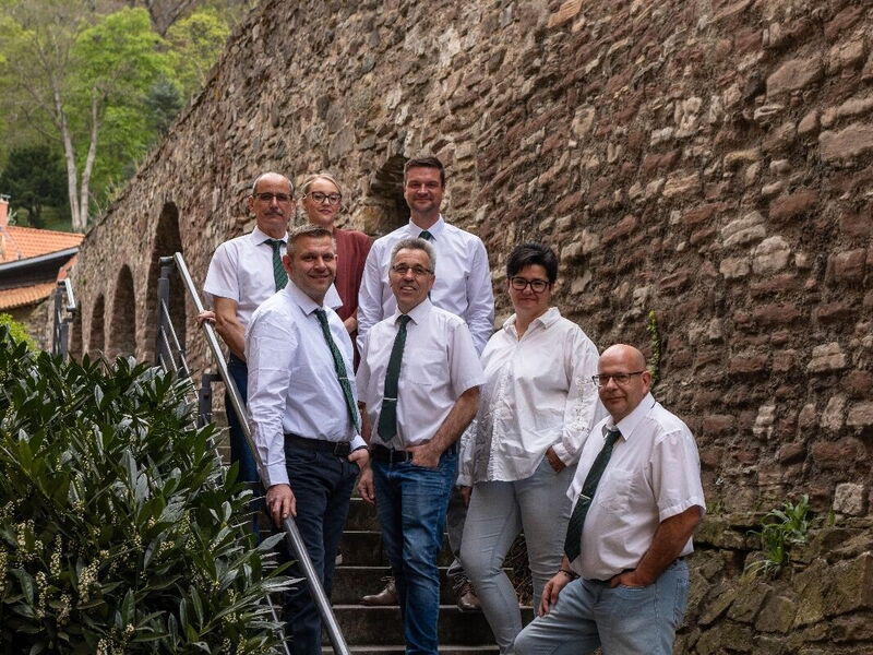 Sieben Personen in weißen Hemden und Krawatten stehen auf einer Steintreppe vor einer alten Steinmauer