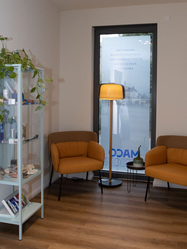 Wartebereich mit zwei orangefarbenen Sesseln, einem Stehlampen und einem Regal mit Pflanzen