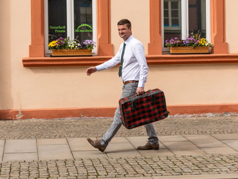 Ein Mann in weißem Hemd und Krawatte geht mit einem karierten Koffer über einen Gehweg