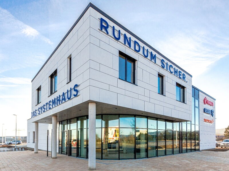 Modernes Gebäude mit der Aufschrift "RUNDUM SICHER" und "DAS SYSTEMHAUS" in blauen Buchstaben, große Glasfront