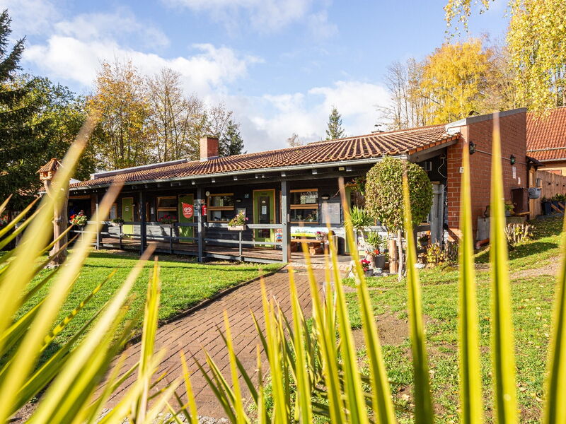 Ein eingeschossiges Gebäude mit dunkler Holzfassade und rotem Ziegeldach, davor ein gepflasterter Weg und