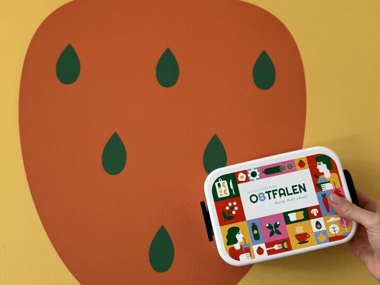 Eine Hand hält eine bunte Brotdose mit der Aufschrift OSTFALEN Kurz mal raus vor einer großen stilisierten