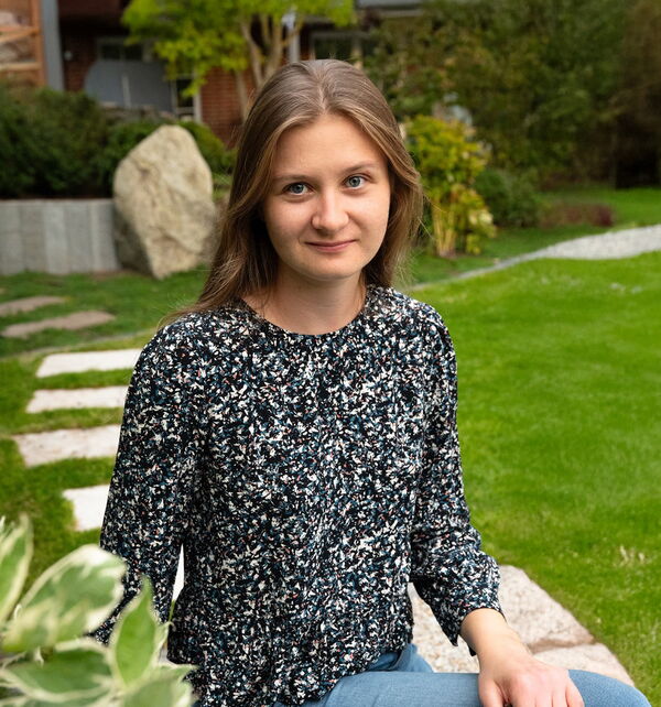Eine junge Frau mit langen braunen Haaren und blauen Augen sitzt im Freien auf einem Stein in einem Garten