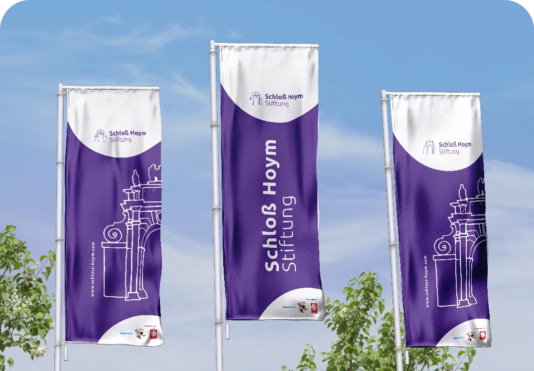 Drei violette Hissfahnen der Schloß Hoym Stiftung mit weißem Logo und Torzeichnung vor blauem Himmel